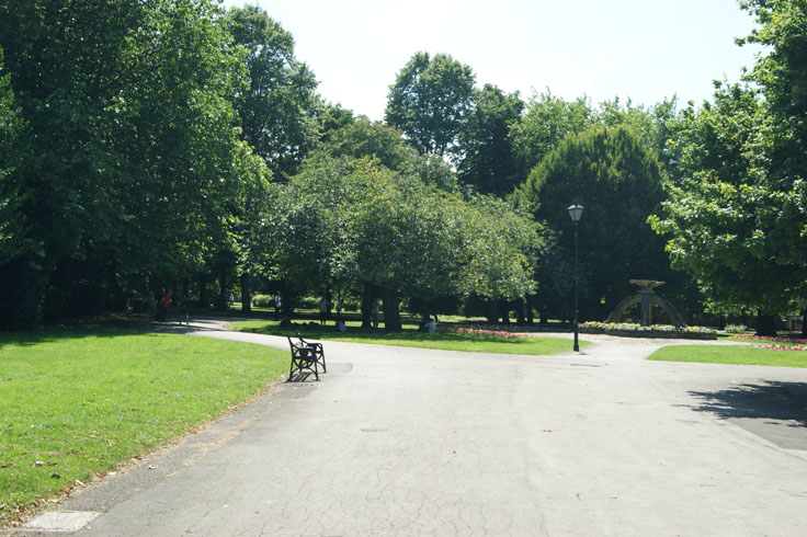 A group of people lying on the grass in a park. In front of them is a flower bed and bench, and on the right a fountain.